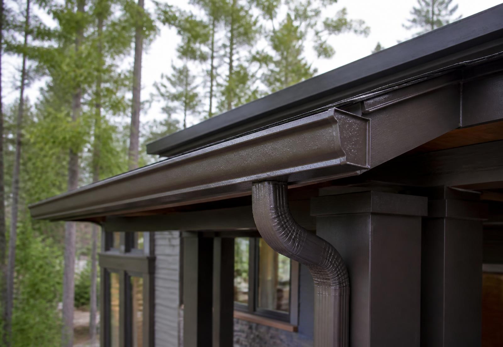Newly installed dark seamless aluminum gutter and downspout along a wooded home's roofline